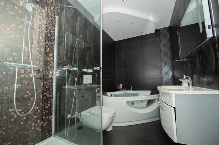Modern bathroom with black tile walls, glass shower enclosure with mosaic accent, white curved bathtub, and white vanity sink.