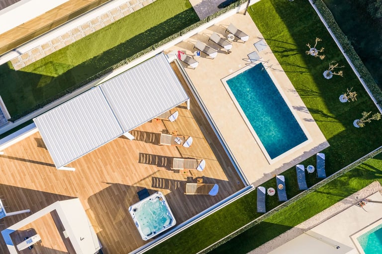Aerial view of a luxury residential pool area with wooden deck, lounge chairs, and blue swimming pools surrounded by manicured green lawn