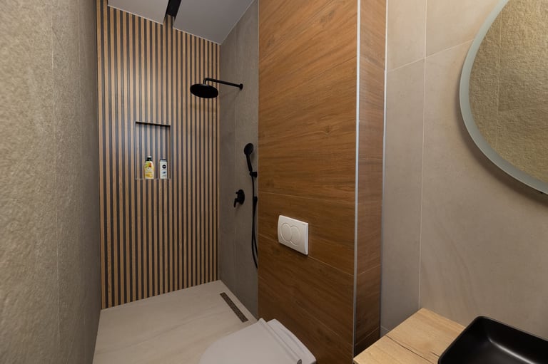 Modern bathroom with walk-in shower featuring vertical slat wood paneling, black fixtures, white base, and round mirror on beige wall