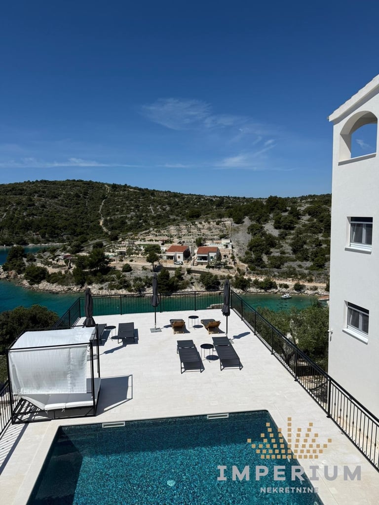 Modern white building with pool deck overlooking turquoise sea and Mediterranean hillside with scattered houses