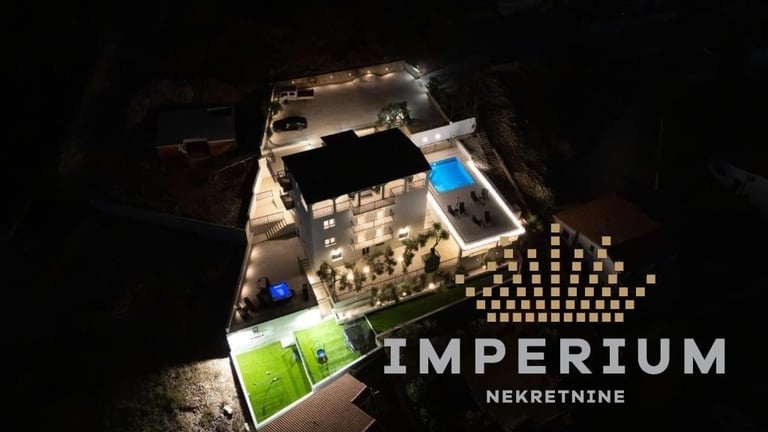 Aerial night view of Imperium Nekretinme building complex with illuminated structures, checkered plaza, and green courtyard area