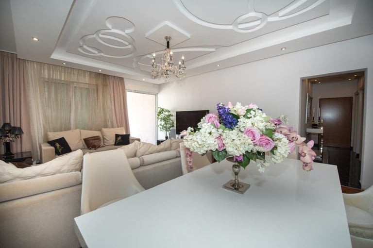 Modern luxury living room with white dining table displaying pink and white flower arrangement, cream sofa, decorative ceiling, and crystal chandelier