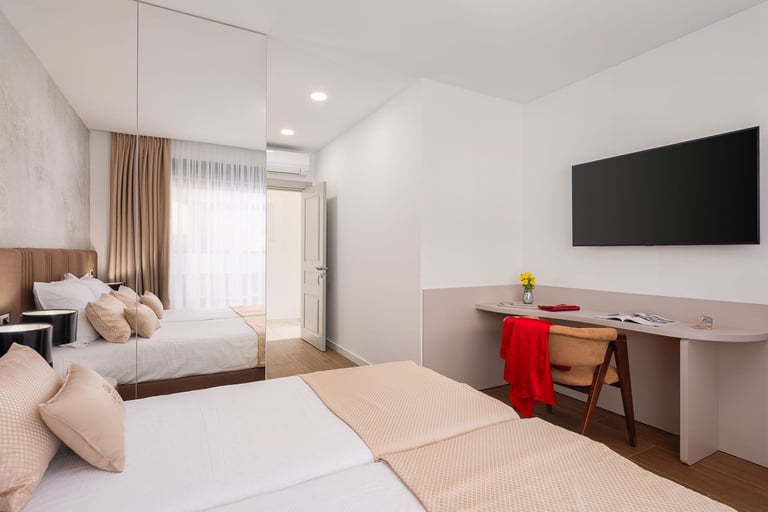Modern hotel room with two beds, desk with TV, and glass door leading to balcony with curtains