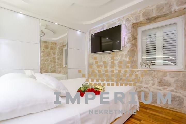 Modern bathroom and bedroom combined space with white bed, glass shower enclosure, and beige stone tile walls with mounted TV