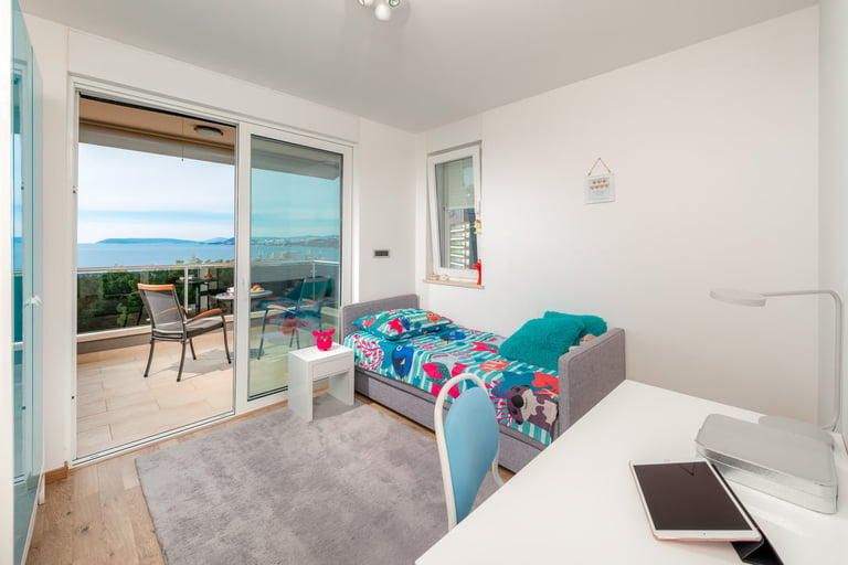 Bright bedroom with blue-patterned bed, desk, and large glass doors opening to a coastal balcony with ocean view