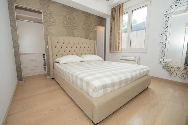 Modern bedroom with beige upholstered bed, wooden flooring, patterned wallpaper, and large windows with natural light