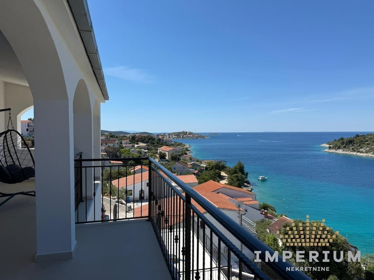 Balcony view of turquoise Mediterranean waters, coastal cliffs, and terracotta-roofed buildings on a sunny day