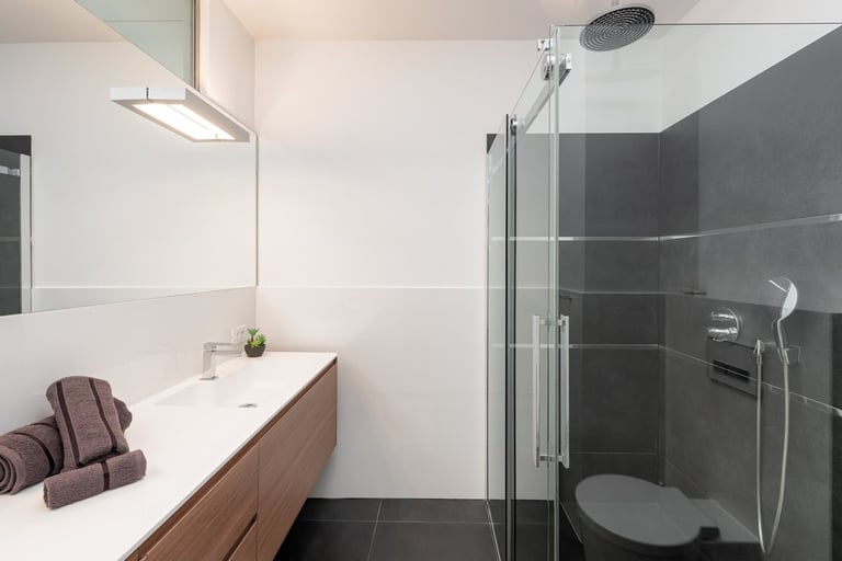 Modern bathroom interior with wood vanity, white sink, glass shower enclosure with dark tile, and rolled towels on counter