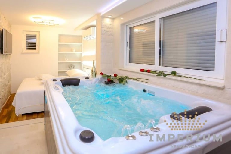 Modern spa bathroom with illuminated hot tub filled with blue water, single bed visible in background, white walls, and large window with blinds