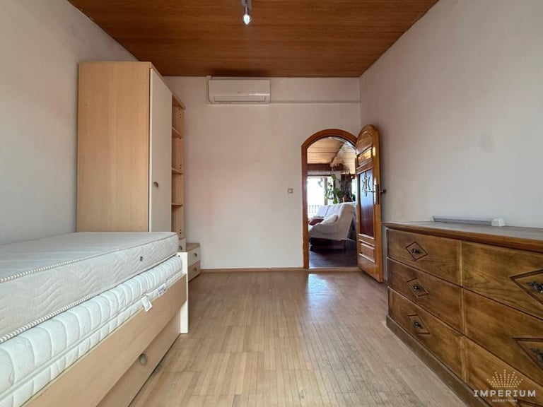 Bright bedroom with wooden ceiling, single bed on left, wooden dresser on right, and open doorway to adjacent room