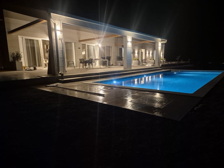Modern minimalist house at night with illuminated interior, featuring a glowing blue swimming pool and dark terrace
