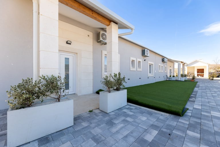 Row of modern white townhouses with paved courtyard, white planters, and artificial green lawn under clear blue sky