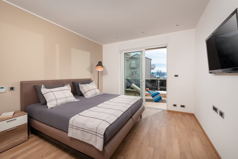 Modern bedroom with a king bed, wooden flooring, wall-mounted TV, and glass doors opening to a balcony with city views