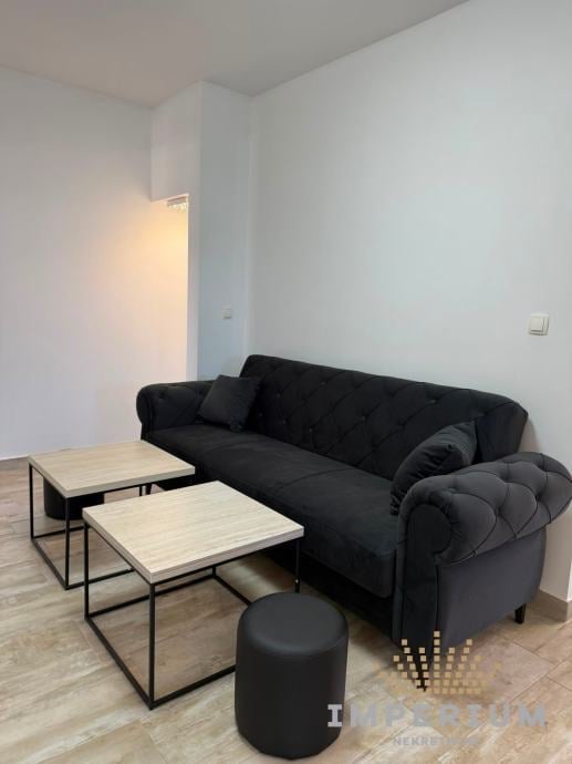Minimalist living room with black sofa, two wooden side tables, and black ottoman in a bright white room