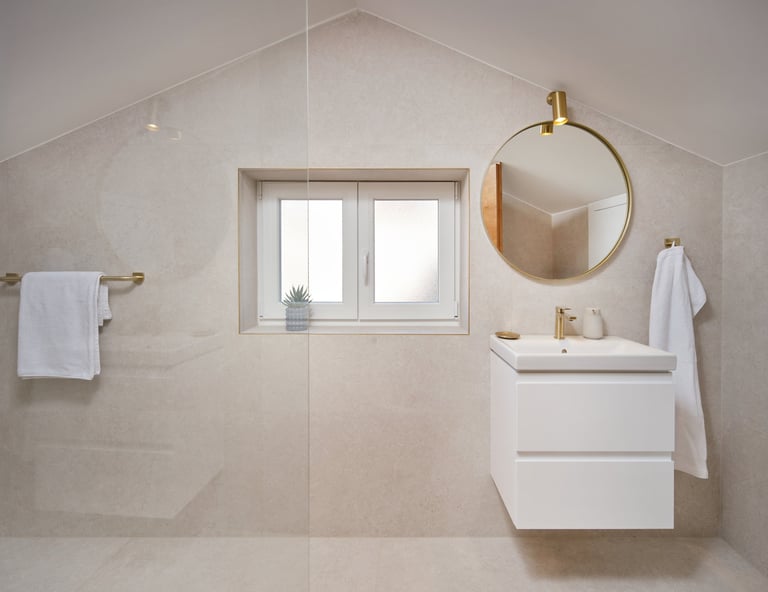 Modern minimalist bathroom with neutral beige tiles, white floating vanity, round gold-framed mirror, and window with natural light