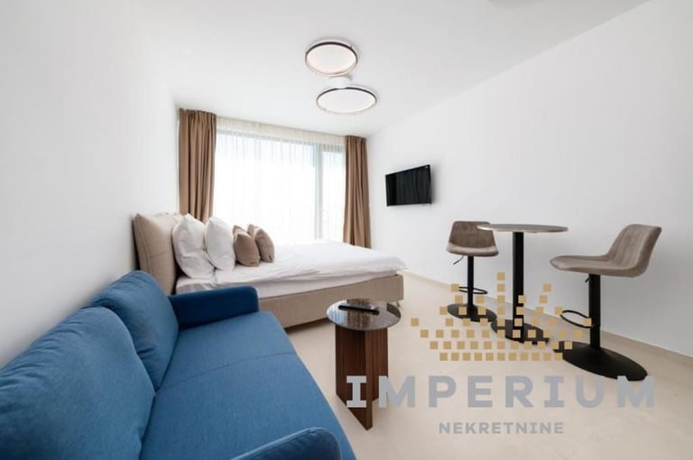 Modern studio apartment with blue sofa, window seating area with cushions, desk with chair, and wall-mounted TV