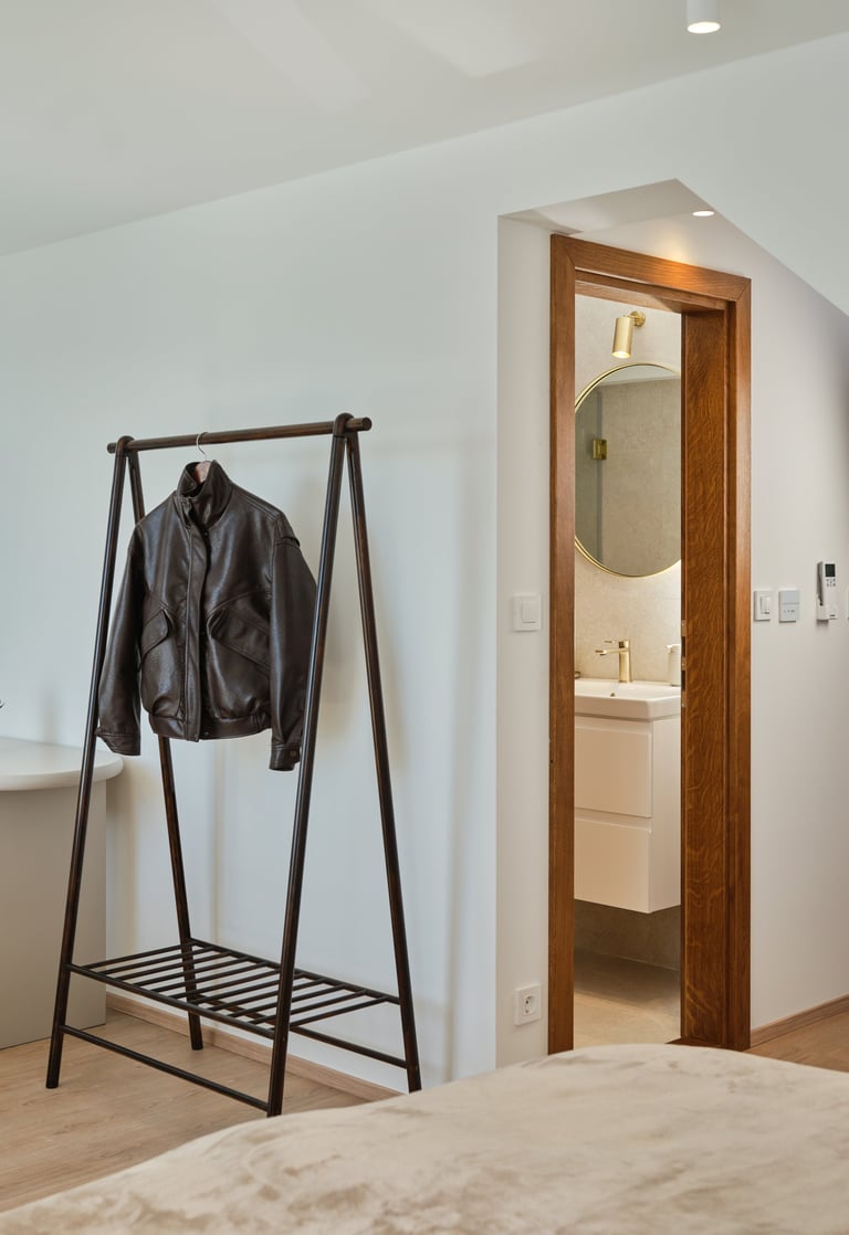 Modern bedroom with black metal clothing rack holding a leather jacket and wooden-framed bathroom doorway with mirror visible