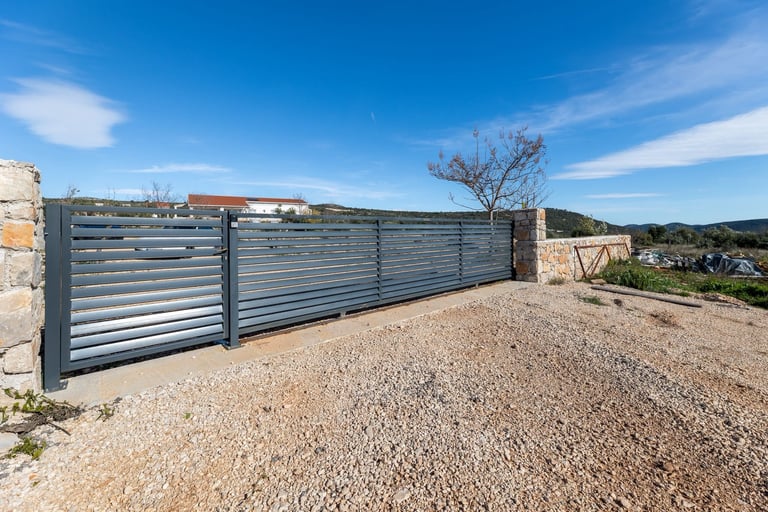 Modern metal sliding gate in driveway with gravel surface, hillside landscape, and clear blue sky