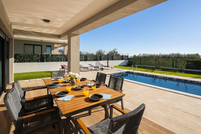Modern patio with wooden dining table, black chairs, pool, manicured lawn, and countryside views under clear skies