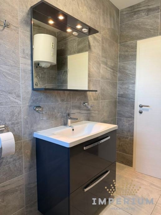Modern bathroom vanity with dark blue cabinet, white sink, illuminated mirror, and gray tile walls
