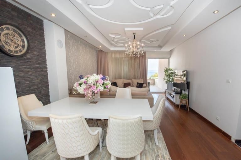 Modern dining room with white oval table, cream quilted chairs, chandelier, dark accent wall, and floral centerpiece arrangement