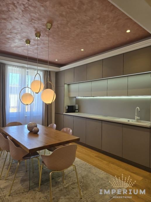 Modern kitchen-dining room with wooden table, beige chairs, pendant lights, and sleek gray cabinetry with built-in shelving and appliances.