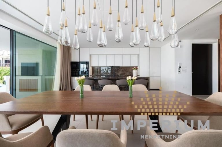 Modern dining room with wood table, beige chairs, hanging pendant lights, and open-concept kitchen in background