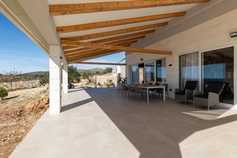 Modern house exterior with covered patio, wooden beams, dining area, and mountain views in background