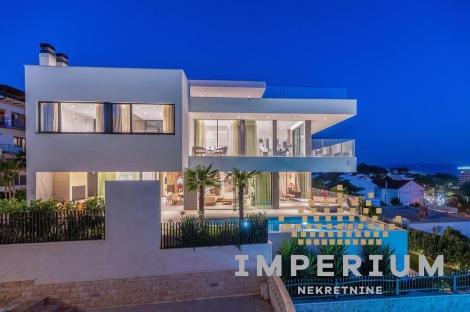 Modern white multi-story house with large windows illuminated at dusk, featuring a pool and mountain views in the background