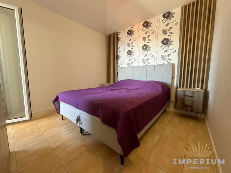 A bedroom with a bed draped in a purple blanket, white headboard with botanical artwork, wooden flooring, and beige walls with curtains