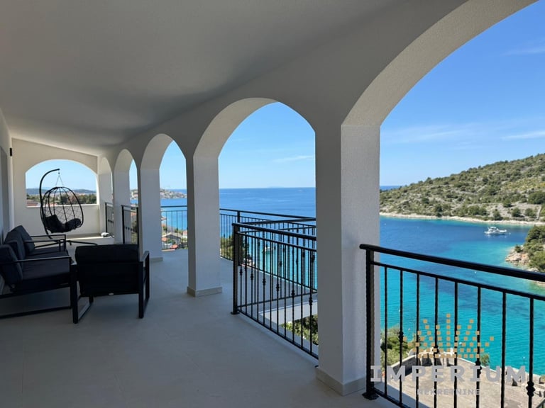 Mediterranean coastal balcony with white arches, comfortable seating, and turquoise sea view with sailboat