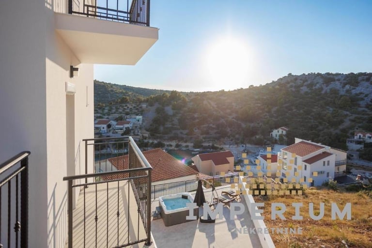 Modern white building balcony overlooking a Mediterranean coastal village with blue swimming pool, terracotta roofs, and forested hillside at sunset