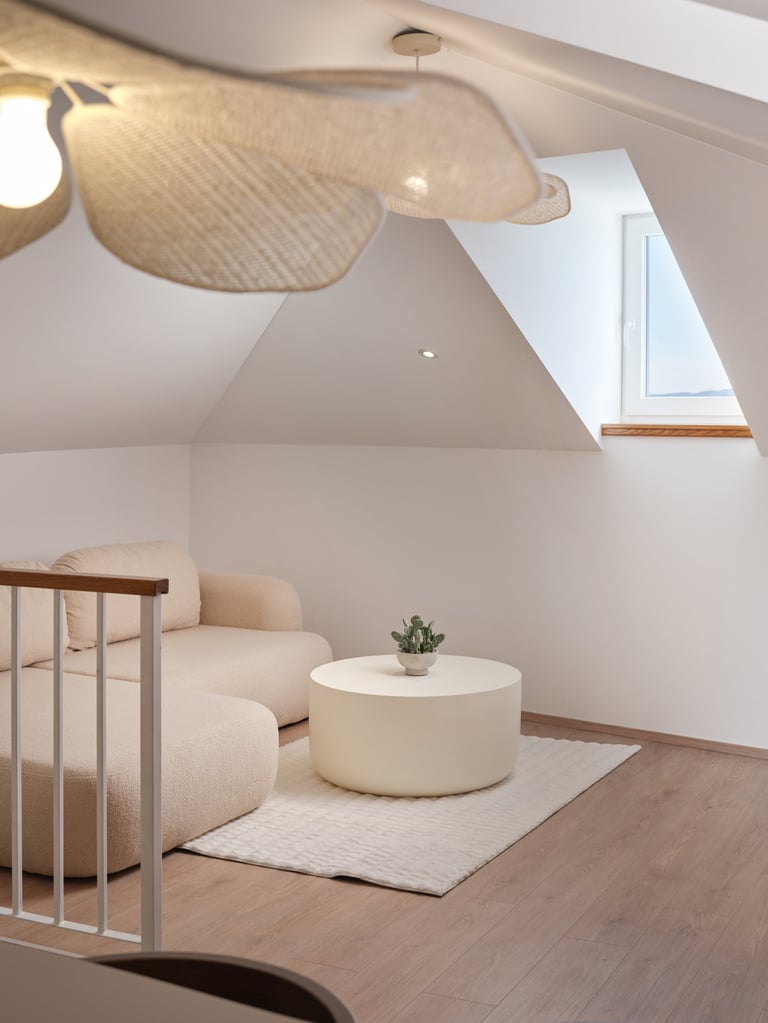 Minimalist attic bedroom with cream armchair, round white ottoman, small potted plant, ceiling fan, and skylight window