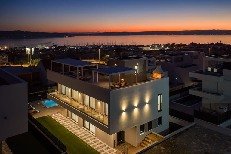 Modern illuminated house with rooftop pool overlooking bay at dusk with city lights in background