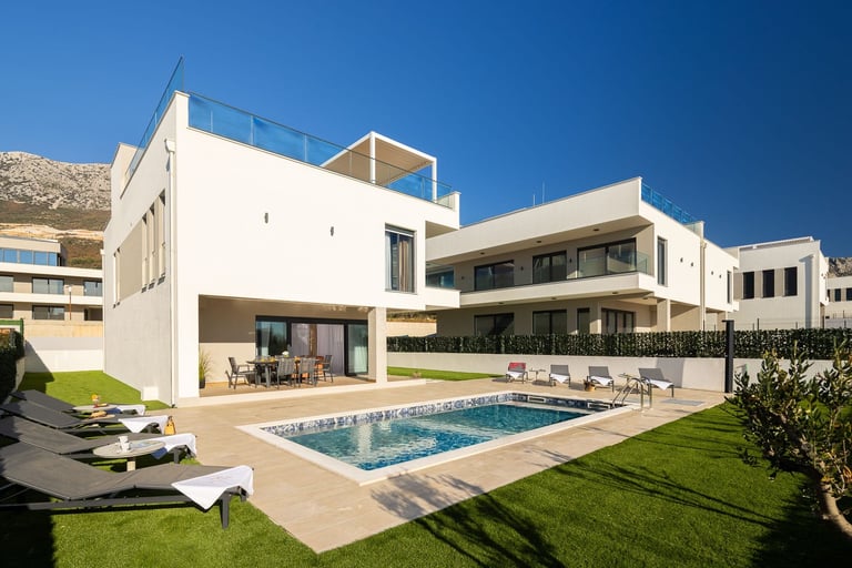 Modern white villa with swimming pool, manicured lawn, lounge chairs, and mountain backdrop under clear blue sky
