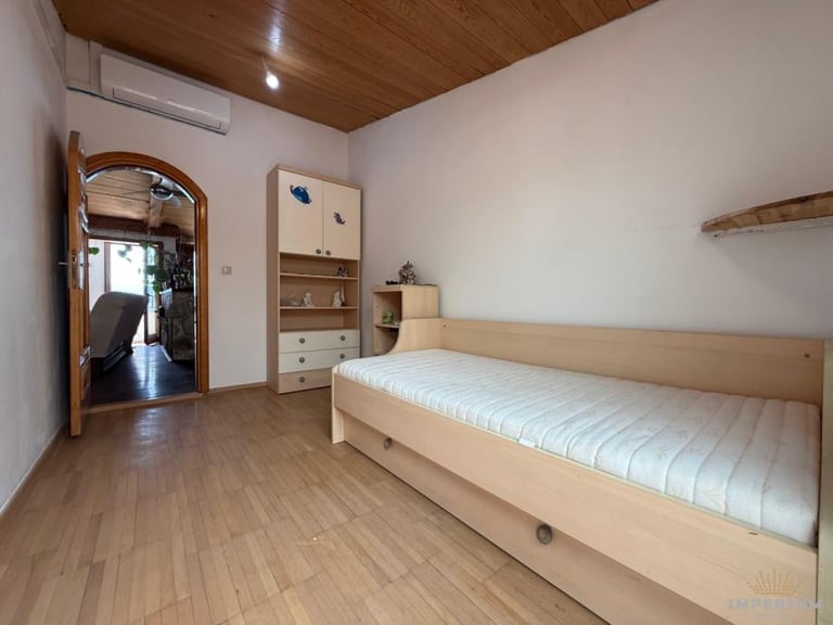 Bright bedroom with wooden ceiling and flooring, featuring a light wood bed with white mattress, shelving unit, mirror, and storage dresser