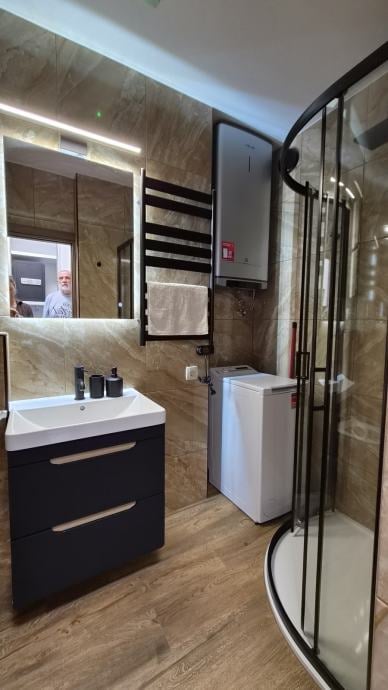Modern bathroom with black vanity, white sink, illuminated mirror, wooden tile flooring, and curved glass shower enclosure