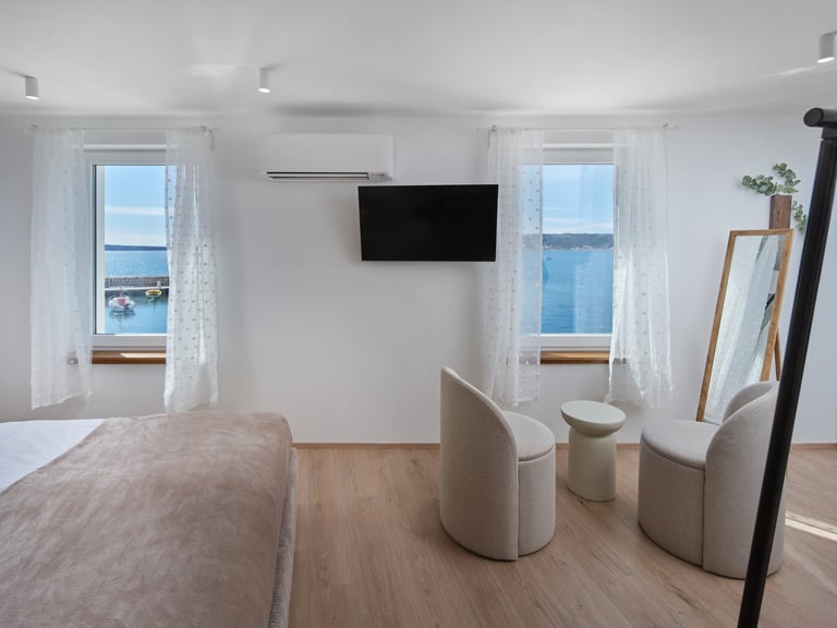 Modern bedroom with ocean view, wall-mounted TV, two white upholstered chairs, wooden flooring, and sheer curtains on windows overlooking the sea