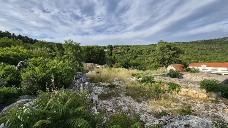 Scenic landscape with green hills, rocky terrain, and a red-roofed house nestled among vegetation under a blue sky