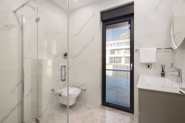 Modern bathroom with marble walls, glass shower enclosure, wall-mounted toilet and sink, and a window overlooking a building