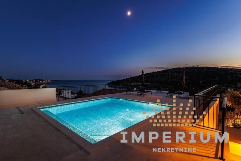 Illuminated swimming pool at Imperium hotel overlooking a coastal town at twilight with moon visible in sky