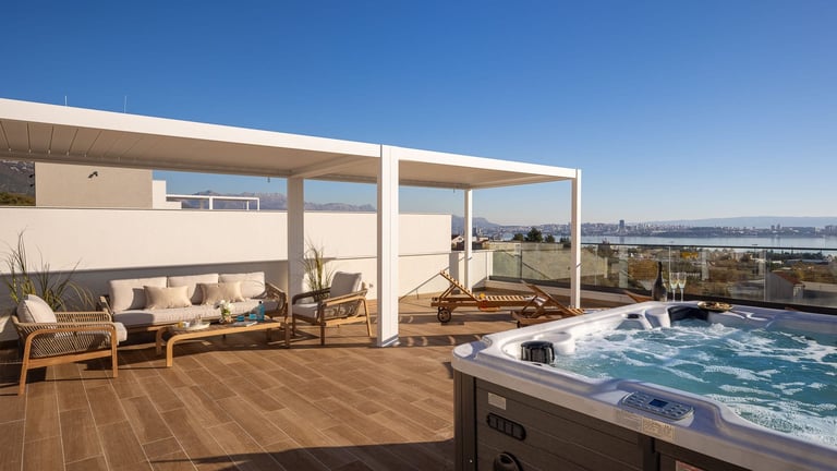 Modern rooftop terrace with hot tub, wooden deck, lounge seating, and panoramic city waterfront views under clear blue sky