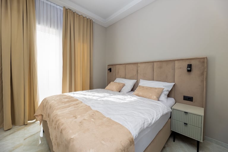 Modern bedroom with a queen bed, beige headboard, cream curtains, nightstand, and white walls