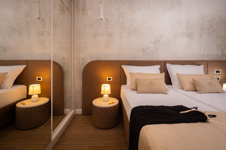 Modern hotel bedroom with large mirror, queen bed with white linens, brown accent nightstands with lamps, and wooden flooring