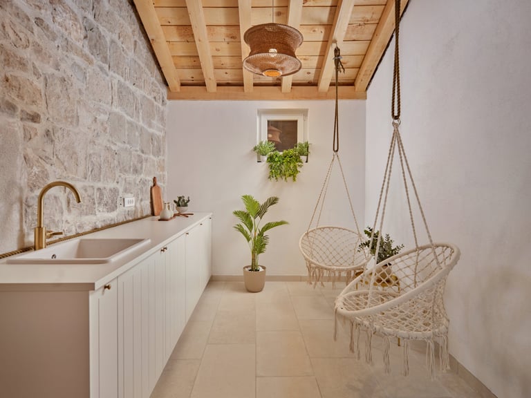 Modern kitchen and relaxation space with white cabinetry, hanging macramé chairs, potted plants, and wooden ceiling fixtures