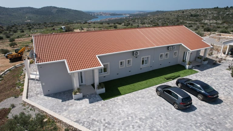 Modern white house with terracotta roof and paved driveway, two cars parked outside, overlooking Mediterranean coastline and hills