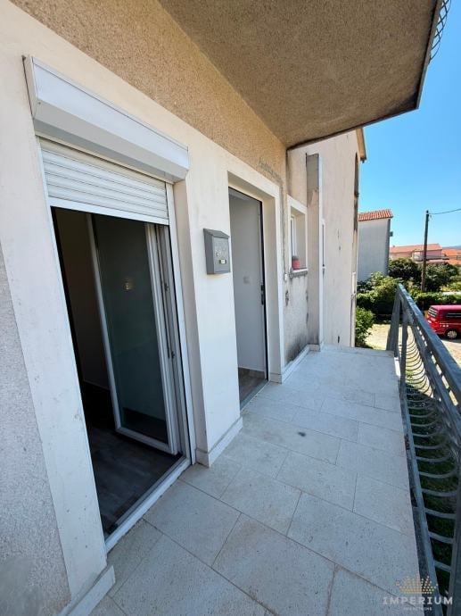 Exterior balcony or terrace with tiled flooring, sliding glass door, and white-framed entrance doors under an overhanging roof