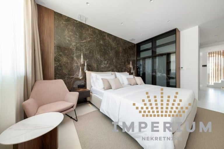 Modern bedroom with textured dark accent wall, white bed with patterned throw, pink armchair, and dark glass wardrobe doors