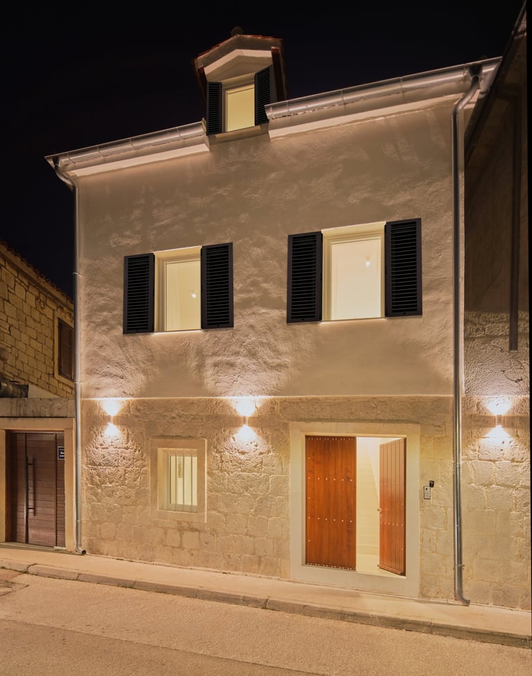 Nighttime exterior of a two-story stone house with illuminated facade, dark wooden shutters, and a red-orange front door