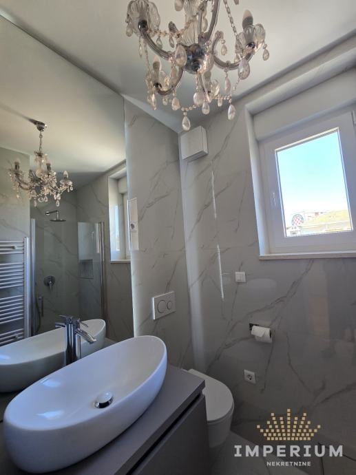 Modern luxury bathroom with white oval sink, crystal chandelier, marble walls, heated towel rail, and bright window overlooking landscape